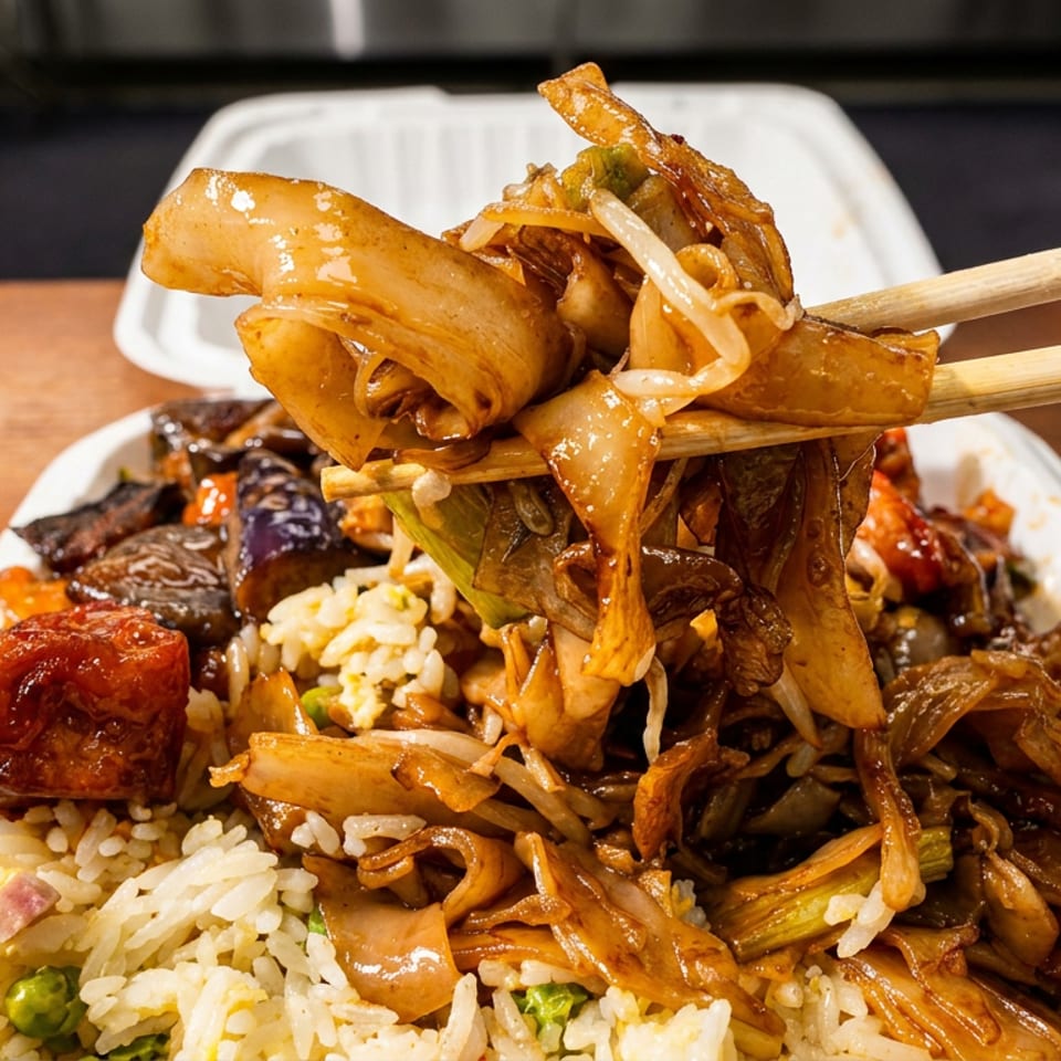 Chopsticks lift saucy stir-fried noodles and vegetables above a takeout platter of fried rice and assorted cooked dishes.